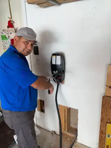 An electrician from Intel Power performing an EV charger installation in a residential garage in Coquitlam. The technician is wearing a blue uniform and a gray hat, focusing on wiring the EV charging unit securely to the wall.